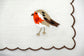 White linen cocktail napkin with orange brown bird embroidery.