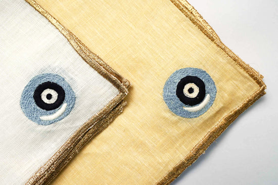 White linen cocktail napkin with round blue evil eye embroidery.